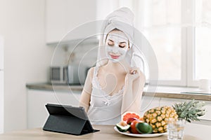 Portrait of young pretty woman with mud facial mask on her face and hair wrapped in towel, sitting at the table with