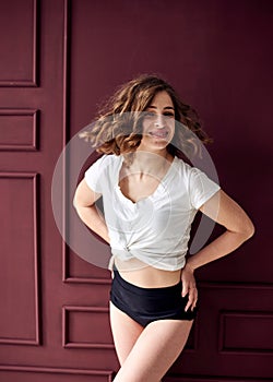 A portrait of young pretty woman with short curly hair, wearing white top and black shorts, standing by burgundy wall, with hands