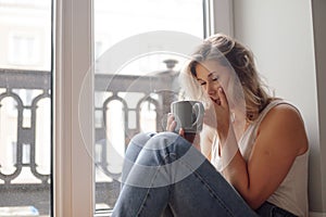 Portrait of young pretty woman sit with closed eyes on floor near window, holding grey cup, covering mouth with hand.