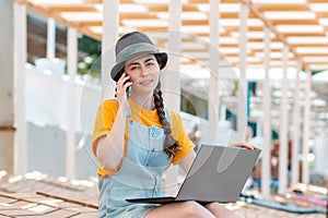 Portrait of young pretty woman is sitting on a sunbed on the beach with a laptop and talking on the phone. In the background beach