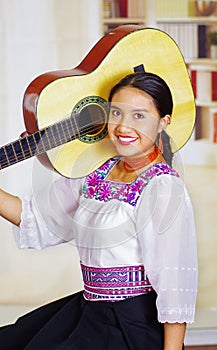 Portrait of young pretty woman wearing beautiful traditional andean clothing, sitting down holding acoustic guitar up