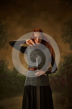 Portrait of young princess in chainmail with sword posing against vintage background, showing her brave and beauty.