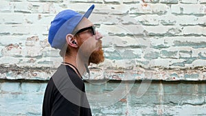 Portrait of young red bearded man in black t-shirt and snapback in rock style on urban background listening to music headphones