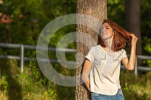 Portrait of a young red-haired girl. The girl leaned against a tree and enjoys the evening sun during sunset