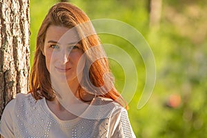Portrait of a young red-haired girl. The girl leaned against a tree and enjoys the evening sun during sunset
