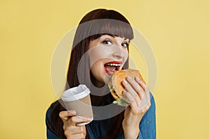 Portrait of young red haired hungry woman holding take away coffee cup and biting fresh hamburger, smiling to camera
