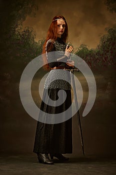 Portrait of young red haired princess in chainmail with sword posing against vintage background, showing her brave and