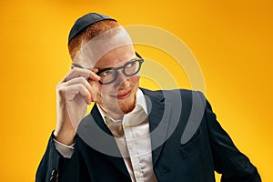 Portrait of young redhead Jewish man in yarmulke, glasses and suit smiling, posing against yellow studio background