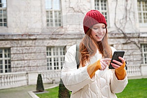 Portrait of young redhead woman looking at her mobile phone, checks her location on smartphone app, walking around city