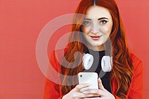 portrait of young redhead woman smiling, with elements of technology, wireless headphones, and smartphone on red