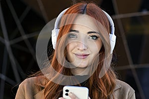 portrait of young redhead woman smiling, with wireless headphones, and smartphone