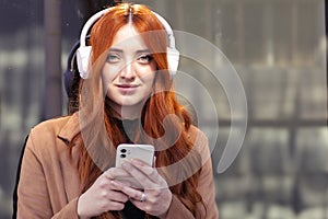 portrait of young redhead woman smiling, with wireless headphones, and smartphone