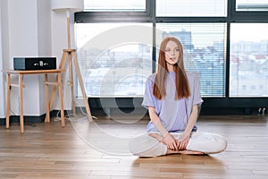 Portrait of young relaxed woman meditating sitting on floor in lotus position on background of window in bright office