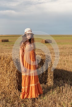 Portrait of a young rural woman in a sarafan and hat that stands near a haystack in a wheat field with a stormy sky