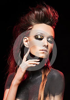 Portrait of a young savage girl. Naked shoulders and neck are covered with black paint. Conceptual makeup with gold leaf on her