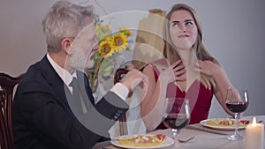 Portrait of young seductive woman in red dress sitting with senior man at the table and smiling. Charming woman of easy