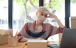 A portrait of young serious Asian woman working with laptop in the office full of packages and boxes stacking up, busy