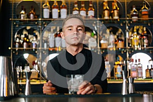 Portrait of young serious bartender standing behind bar counter and holding stirring spoon and glass filled with ice