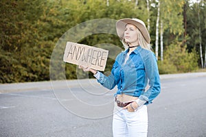 Portrait of young serious blond woman raising cardboard with inscription anywhere, peering looking narrowly. Travelling.