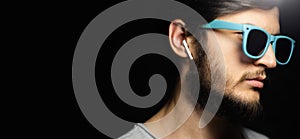 Portrait of young serious man with wireless earphones, looking up, wearing blue sunglasses, isolated on black background.