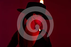 Portrait of young sexy woman in hat on dark red studio background with infrared light. Femme fatale, stylish outfit of