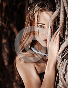 Portrait of young sexy woman savage standing at, hiding behind branches of tropical tree, lianas. Summer vibes