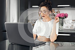 Portrait of a young shocked woman in glasses sitting in front of a laptop computer, looking at the screen. Funny facial