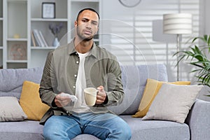 Portrait of a young sick African-American man looking tiredly at the camera. Sitting at home on the couch and holding a