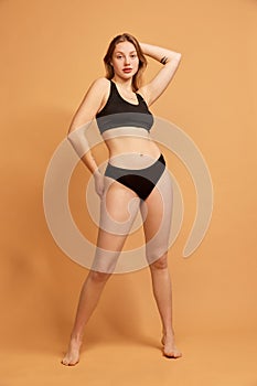 Portrait of young slim beautiful girl wearing black underwear posing over sand color background. Concept of beauty, body