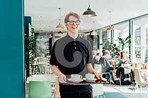 Portrait of young smiling affable waiter carrying on tray with coffee cups to client table in cafe. Hospitality service