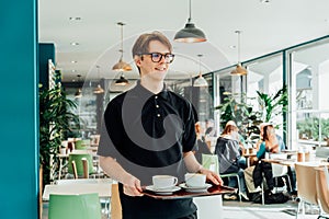 Portrait of young smiling affable waiter carrying on tray with coffee cups to client table in cafe. Hospitality service