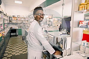 Portrait of young smiling African male pharmacist working with computer behind counter in pharmacy