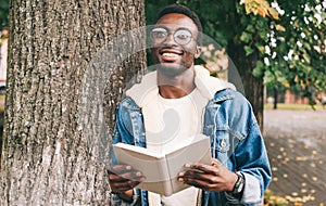 Portrait young smiling african man student with a book looking away wearing an eyeglasses in autumn city park