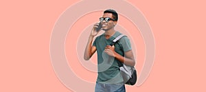 Portrait of young smiling african man student calling on smartphone wearing backpack, sunglasses isolated on pink background