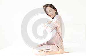 Portrait of a young smiling Asian girl in sweater pink hugging a pillow sitting in a bed at her bedroom. Good morning, new day,