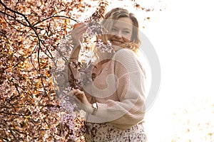 Portrait of young smiling beautiful woman with fair hair wearing pink cardigan, dress standing near bird-cherry tree.