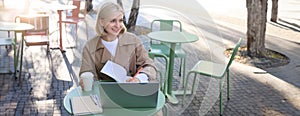 Portrait of young smiling, beautiful woman, studying outdoors in cafe, has laptop and coffee on table, writing in