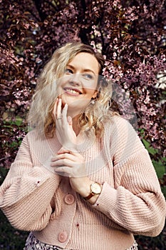 Portrait of young smiling beautiful woman wearing dress, pink cardigan, standing near bird-cherry tree, posing in park.