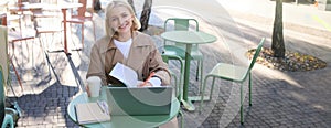 Portrait of young smiling blond woman, working on laptop, sitting in outdoor cafe on street, drinking coffee