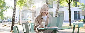 Portrait of young smiling blond woman, working on laptop, sitting in outdoor cafe on street, drinking coffee