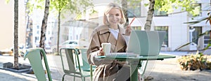 Portrait of young smiling blond woman, working on laptop, sitting in outdoor cafe on street, drinking coffee