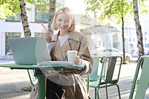 Portrait of young smiling blond woman, working on laptop, sitting in outdoor cafe on street, drinking coffee