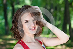 Portrait of young smiling brunette girl in red dress posing on alley in summer park on background of trees