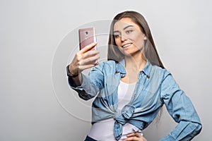 Portrait of a young smiling brunette woman in casual clothes making a selfie with her pink phone against white background.