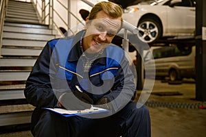 Portrait of a young smiling Caucasian auto mechanic sitting on stairs in the repair shop and looking at camera while writing