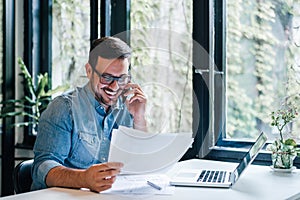 Portrait of young smiling cheerful entrepreneur in casual office making phone call while working with charts and graphs looking at photo