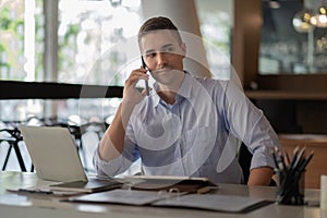 Portrait of young smiling cheerful entrepreneur in modern office making phone call while working with laptop.