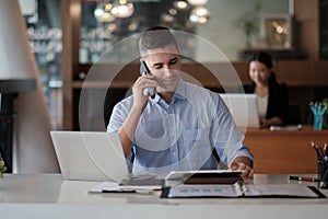 Portrait of young smiling cheerful entrepreneur in modern office making phone call while working with laptop.