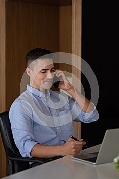 Portrait of young smiling cheerful entrepreneur in modern office making phone call while working with laptop.