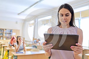 Portrait of young smiling confident female teacher with digital tablet in classroom